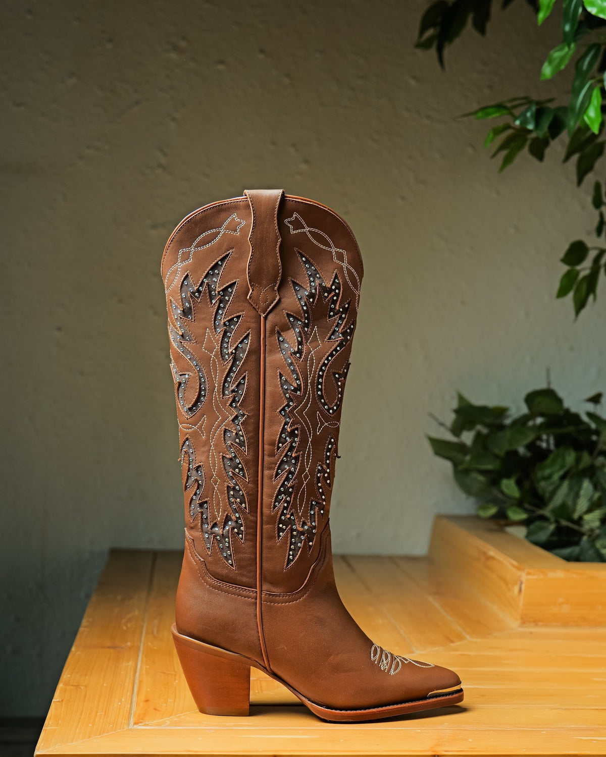 A tall tan Western boot with a stitched shaft on a wooden plinth
