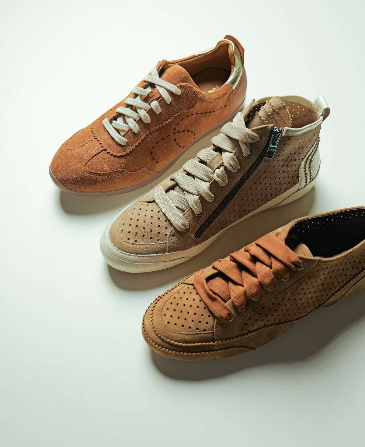 Three women’s perforated suede sneakers in tan, sand and cognac on a light table