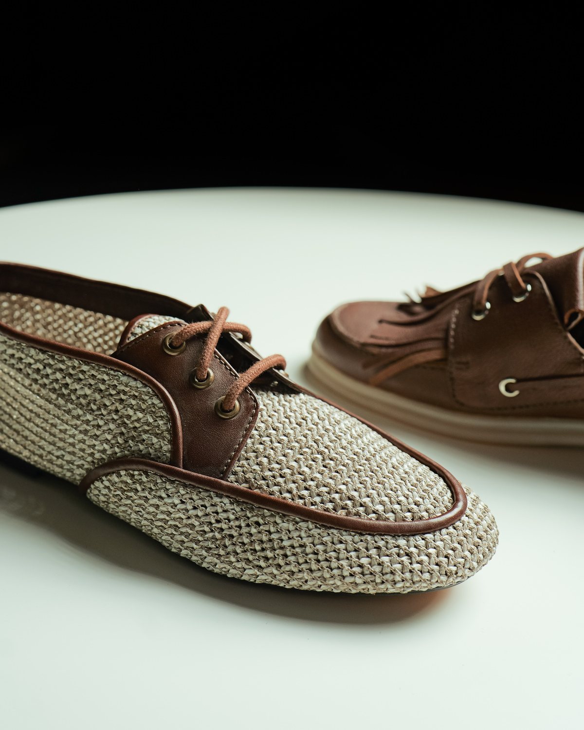 A woven raffia shoe with leather trims beside a leather boat shoe