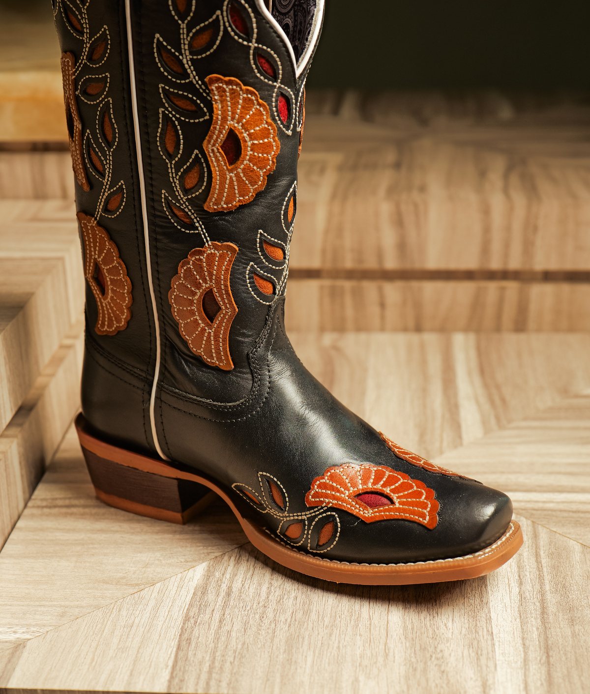 A black cowboy boot with orange floral inlays on shaft, vamp and toe, on a stone plinth
