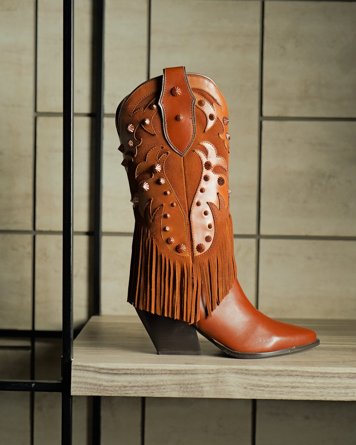 A cognac leather women’s Western-style boot with studs and fringe on a showroom shelf