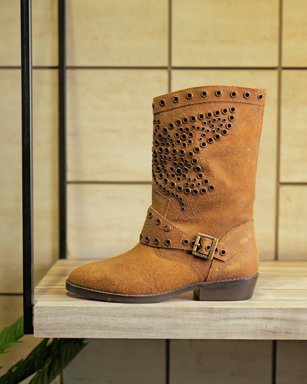 A tan suede women’s mid-calf boot with studded shaft and buckle strap on a shelf